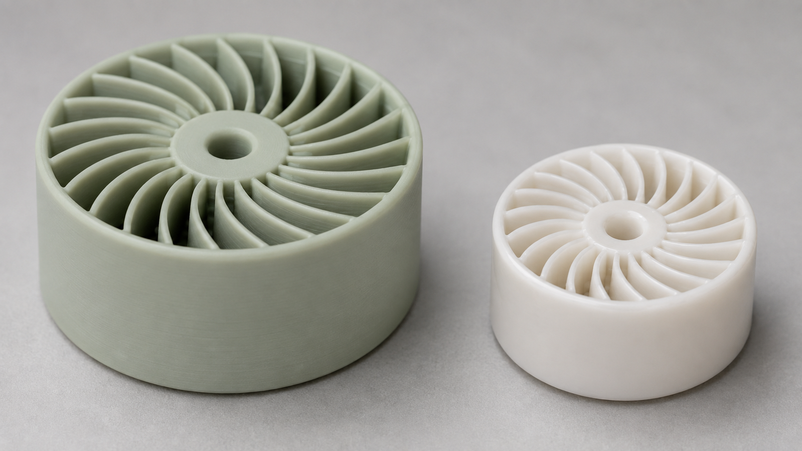 A white alumina ceramic turbine-like part shown in two states side by side on a light studio bench: a larger matte green part fresh from the 3D printer and a smaller, denser, slightly glossy sintered version of the same geometry, illustrating sintering shrinkage