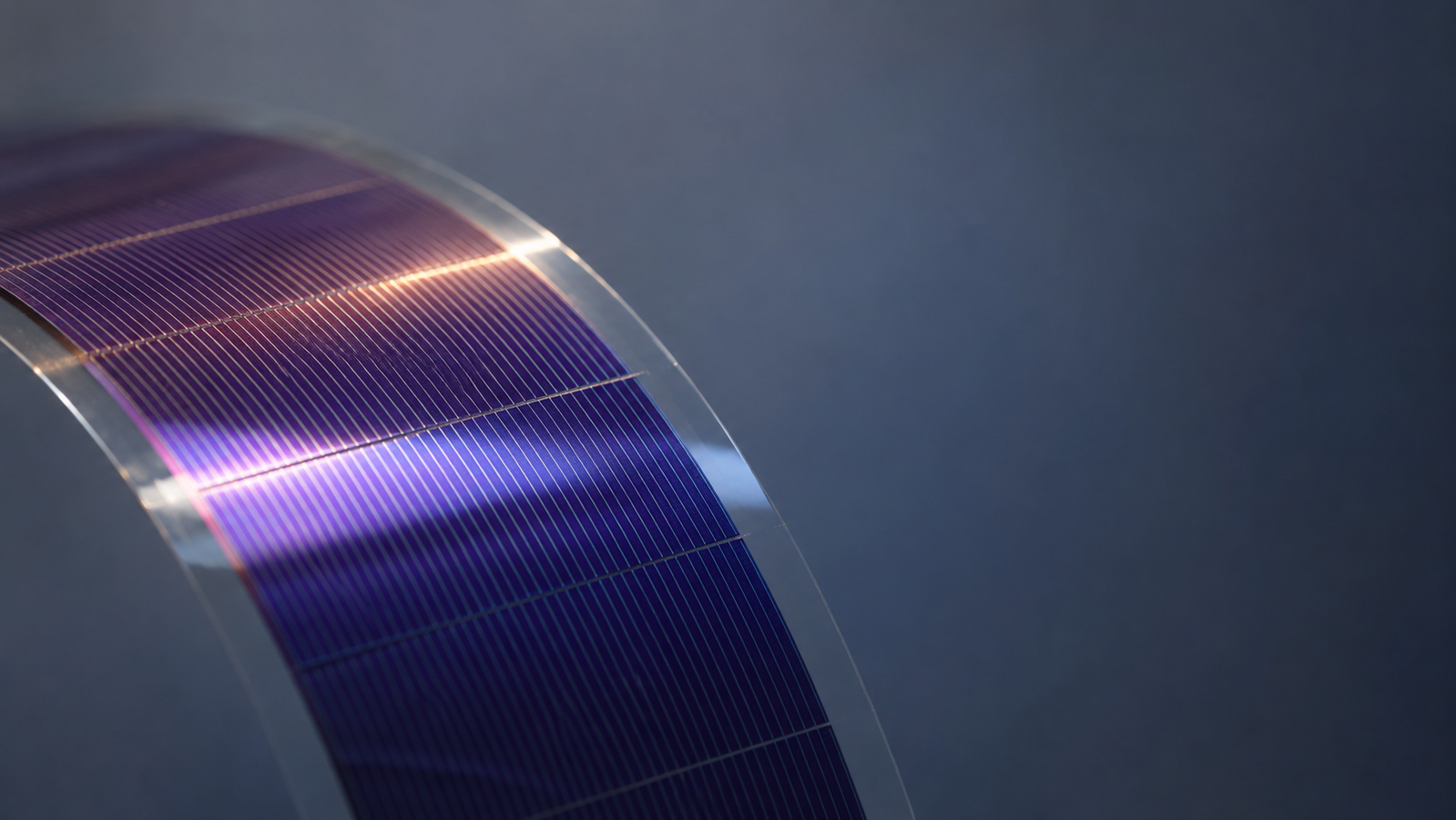 A thin, flexible organic solar cell being gently bent between two hands, with a semi-transparent graphene electrode layer highlighted and sunlight passing through it onto the printed photoactive film beneath