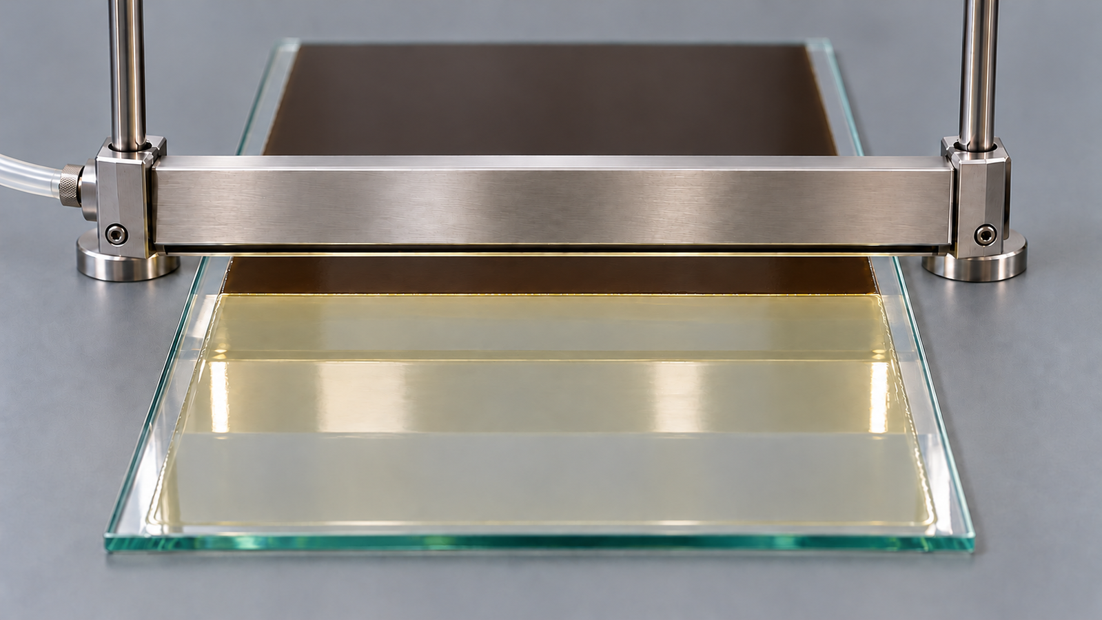 A plain stainless steel air-knife nozzle held on two posts just above a glass plate, with a pale yellow wet film in front of it and a dark, uniform dry film where it has already passed