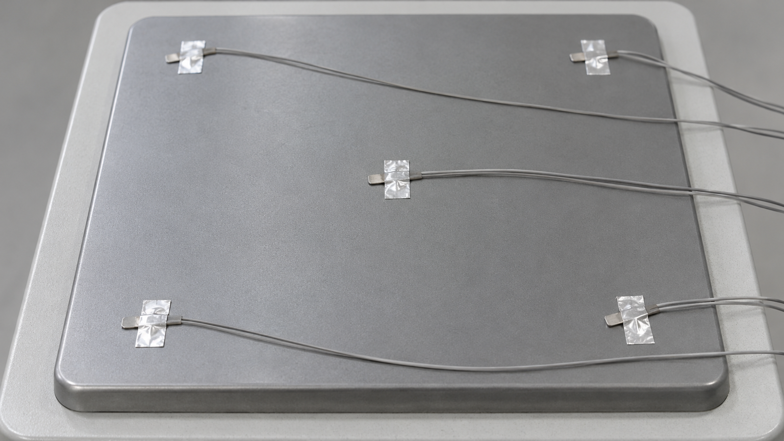 A square matte grey hot plate with thin thermocouple wires taped at several points across its surface